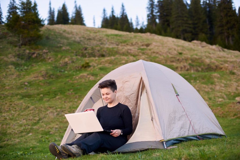 Frau am Laptop neben dem Zelt, als Beispiel für Unterhaltung und Freizeitgestaltung in der Natur.