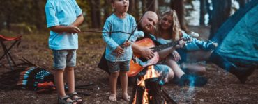 Vier Kinder sitzen lachend um ein Lagerfeuer, grillen Würstchen und sind von einem Zelt und grüner Natur umgeben.