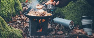 Niewielki piecyk Pocket Stove z gotującym się kubkiem, umieszczony na brzegu jeziora, z nożem obok – doskonały zestaw na outdoor.