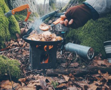 Niewielki piecyk Pocket Stove z gotującym się kubkiem, umieszczony na brzegu jeziora, z nożem obok – doskonały zestaw na outdoor.