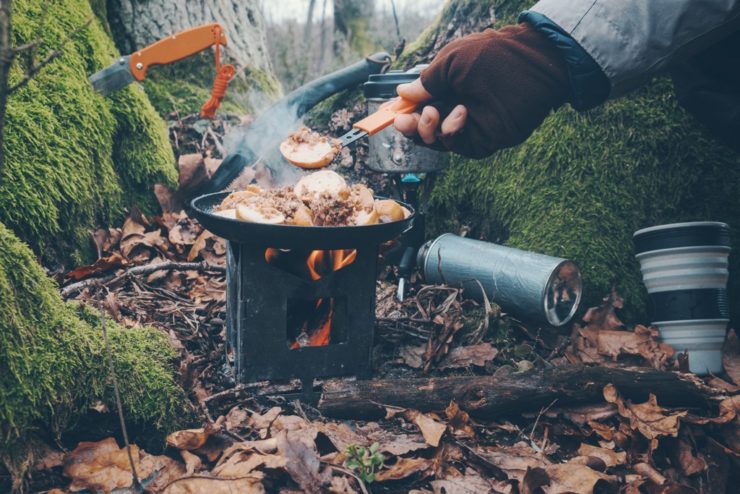 Niewielki piecyk Pocket Stove z gotującym się kubkiem, umieszczony na brzegu jeziora, z nożem obok – doskonały zestaw na outdoor.