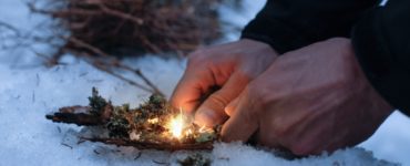 Hände, die Feueranzündmaterial auf Schnee vorbereiten, mit trockenem Moos und einer kleinen Flamme.