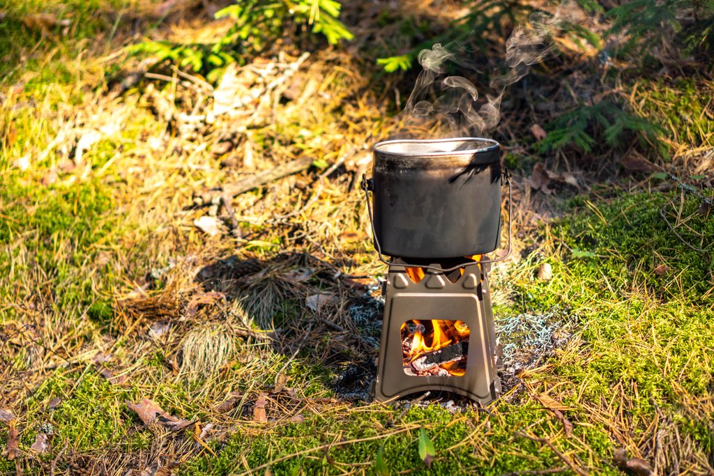 Mały metalowy piecyk Pocket Stove, na którym gotuje się garnek, ustawiony na trawie w otoczeniu natury. W tle widoczna dłoń użytkownika.