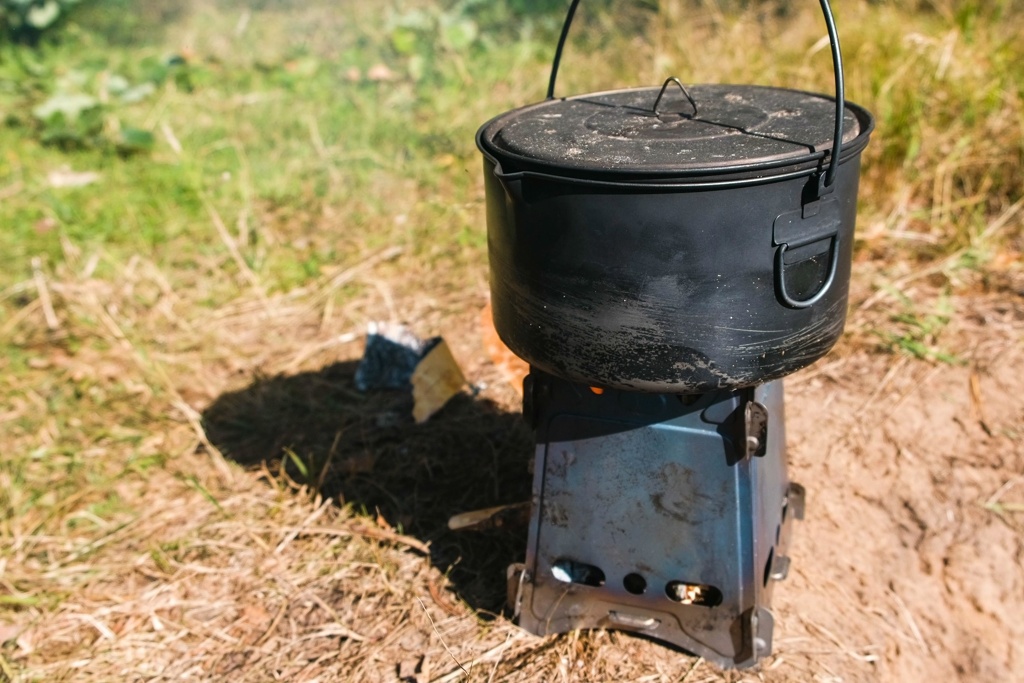 Solidny czarny garnek ustawiony na stalowym piecyku Pocket Stove, umieszczonym na suchym podłożu, idealny na biwak w lesie.