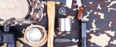 Survival- und Bushcraft-Ausrüstung auf Tarnstoff, darunter ein Beil, ein Klappmesser, ein Seil, eine Tasse, ein Kompass und ein Fernglas.