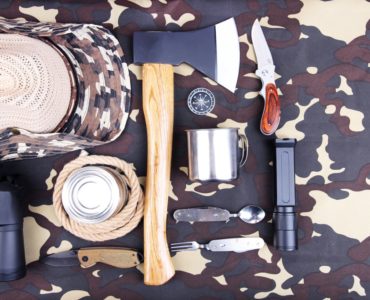 Survival- und Bushcraft-Ausrüstung auf Tarnstoff, darunter ein Beil, ein Klappmesser, ein Seil, eine Tasse, ein Kompass und ein Fernglas.