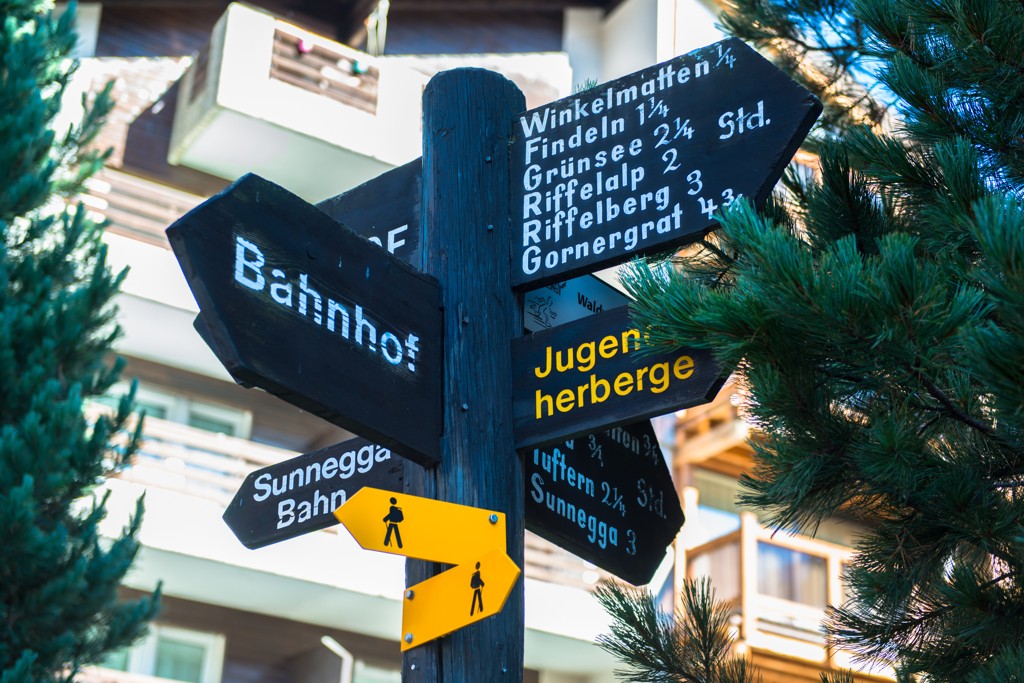 Ein traditioneller schwarzer Holzwegweiser in einem Dorf, mit gelben Wandermarkierungen, die verschiedene Richtungen und Zeiten zu Zielen wie „Bahnhof“ und „Jugendherberge“ angeben.