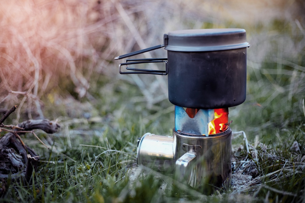 Ein kompakter Kocher mit einer kleinen Flamme erhitzt einen schwarzen Topf in einer grünen Wiese.