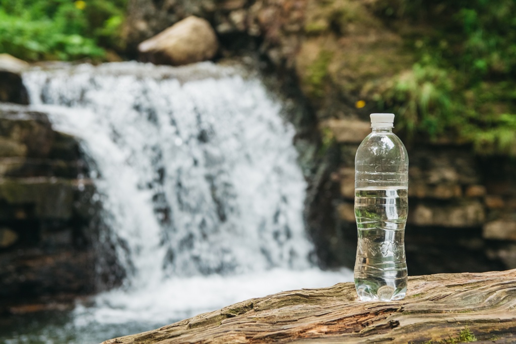 Eine Plastikflasche mit Wasser steht auf einem Holzstamm, vor einem malerischen Wasserfall im Hintergrund.