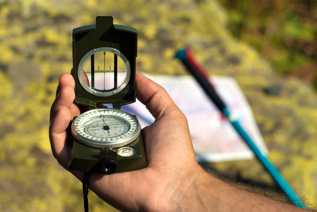 Ein Mann hält einen militärischen Kompass mit integriertem Spiegel und Peillinie in der Hand. Im Hintergrund sind eine Karte und ein Wanderstab zu erkennen.