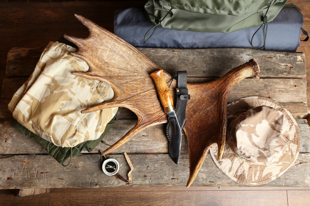 Eine Sammlung von Outdoor-Ausrüstung, darunter ein Geweih, ein Jagdmesser, ein Kompass, Tarnkleidung und ein Hut, arrangiert auf einer rustikalen Holzoberfläche.