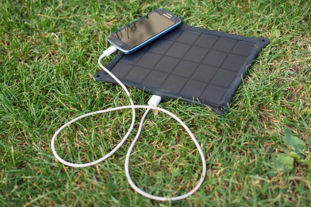 Ein schwarzes, tragbares Solarpanel liegt auf grünem Gras, während es ein angeschlossenes Smartphone auflädt.