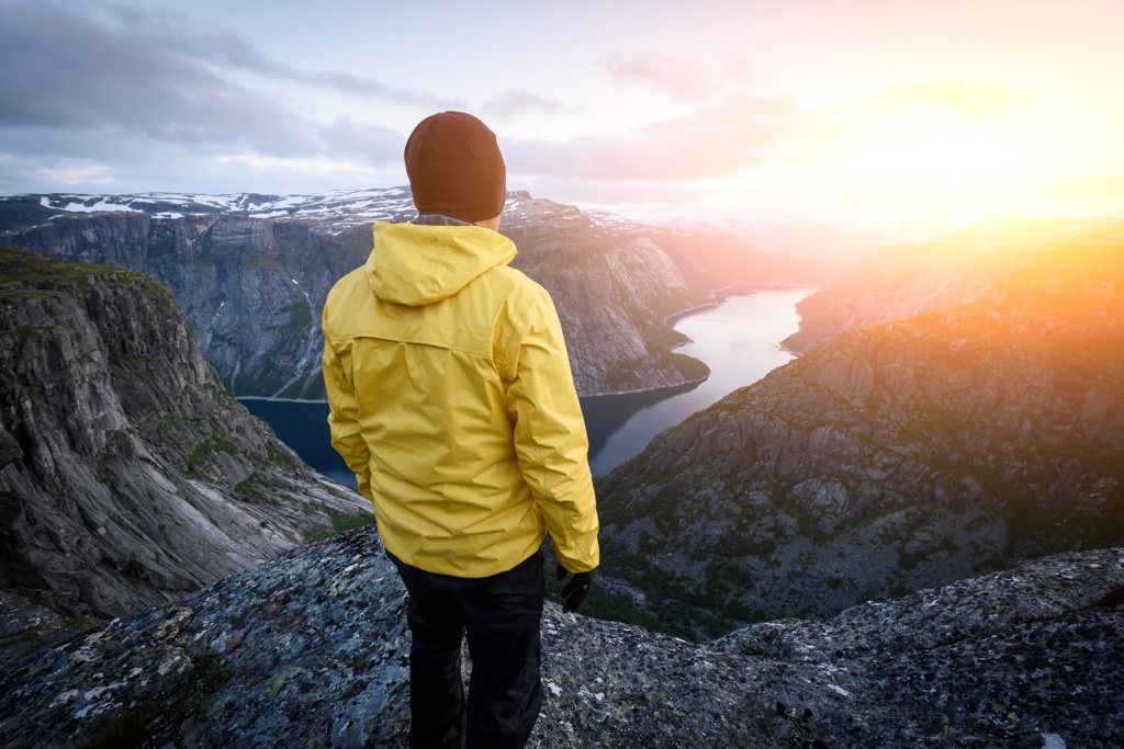 Eine Person in einer leuchtend gelben Jacke betrachtet einen atemberaubenden Sonnenuntergang über einer Berg- und Flusslandschaft.