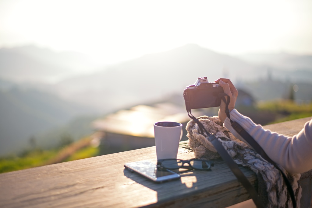 Eine Kamera, Notizbuch, Kaffee und andere Reiseutensilien auf einem Holztisch in einer Berglandschaft.