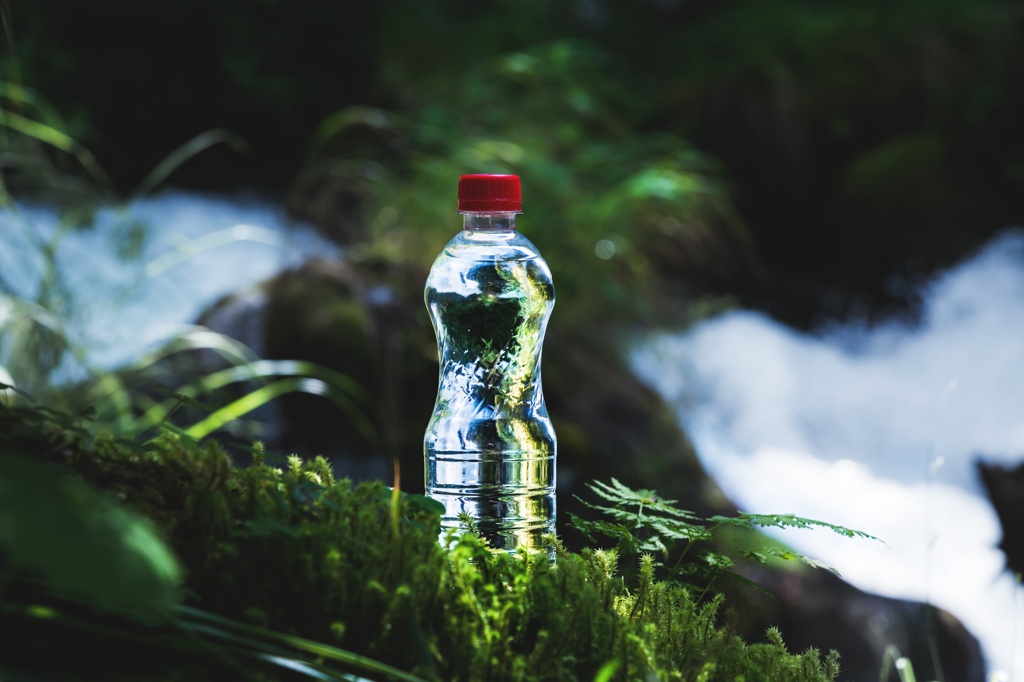 Eine Plastikflasche mit Wasser steht auf grünem Moos, im Hintergrund fließt ein Bach durch den Wald.