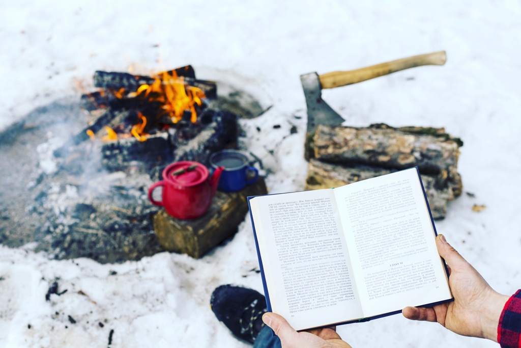 Ein geöffnetes Buch wird vor einem gemütlichen Lagerfeuer gehalten, umgeben von Schnee und einem Beil auf Holzstücken.