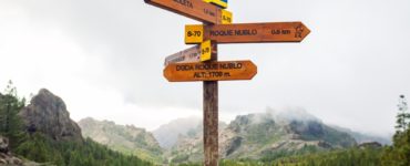 Ein Holzwegweiser in einer bergigen Landschaft mit Schildern, die Ziele wie „Roque Nublo“ anzeigen, begleitet von Symbolen für Wanderer. Im Hintergrund sind Wald und Berge zu sehen.