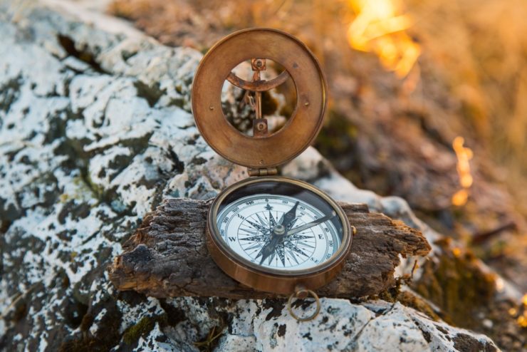 Ein antiker Messingkompass mit geöffnetem Deckel liegt auf einem Stück Holz vor einem felsigen Hintergrund. Der Sonnenuntergang erzeugt ein warmes Licht.