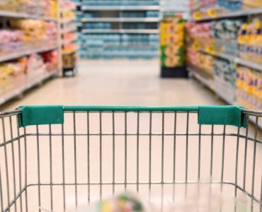 Einkaufswagen mit grünem Griff im Vordergrund, Gänge und Regale voller Waren in einem Supermarkt im Hintergrund.