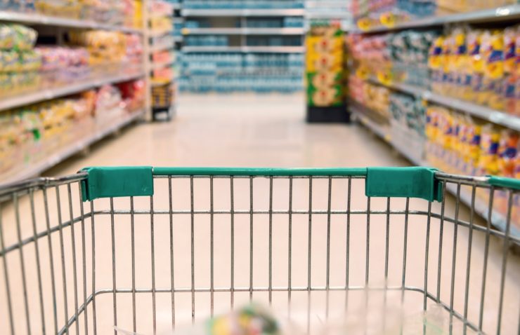 Einkaufswagen mit grünem Griff im Vordergrund, Gänge und Regale voller Waren in einem Supermarkt im Hintergrund.