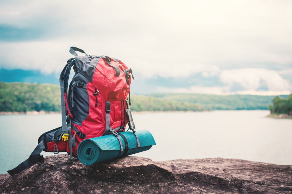 Ein roter Fluchtrucksack mit Isomatte, stehend auf einem Felsen mit Blick auf einen ruhigen See und bewölkten Himmel.