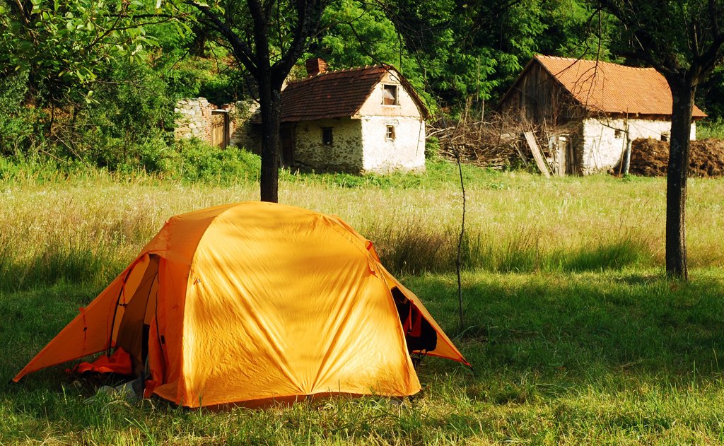 Ein leuchtendes, orangefarbenes Zelt steht auf einer grünen Wiese vor alten, verlassenen Steinhütten.