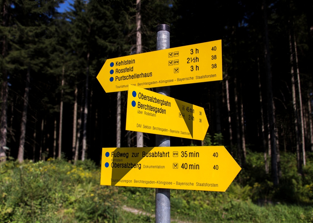 Ein gelber Wegweiser im Wald, der Wanderziele wie „Kehlstein“ und „Rossfeld“ mit Gehzeiten in Stunden und Minuten zeigt. Symbole weisen auf Attraktionen und Wege hin.