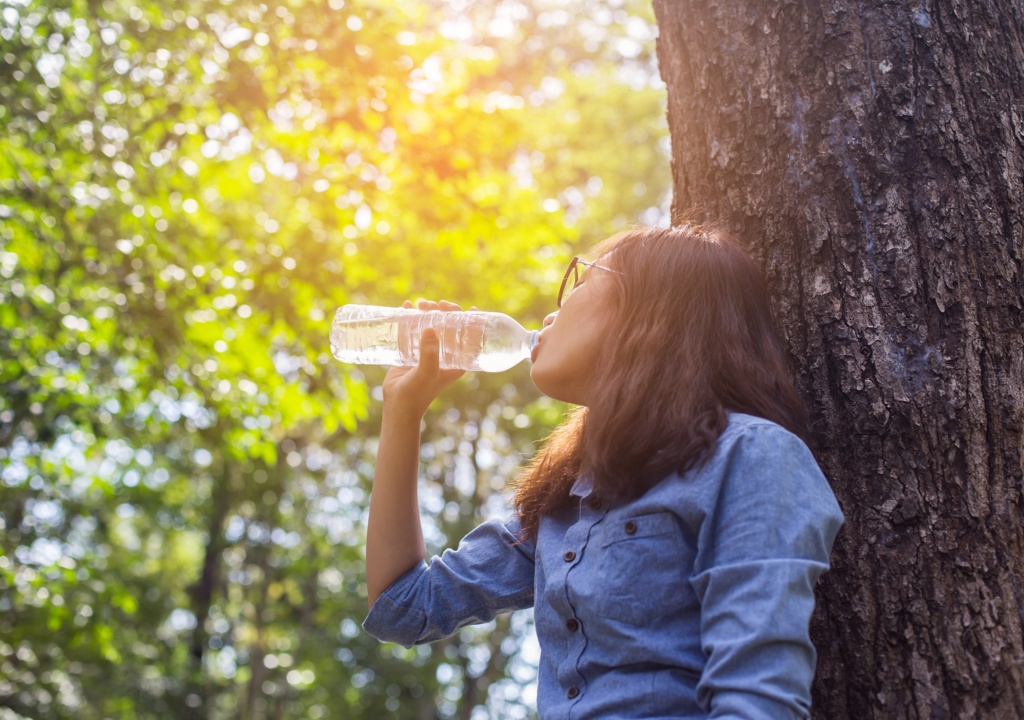 Eine Frau mit Brille lehnt an einem Baum im Wald und trinkt Wasser aus einer Plastikflasche. Sonnenlicht scheint durch die Blätter und schafft eine angenehme Atmosphäre.